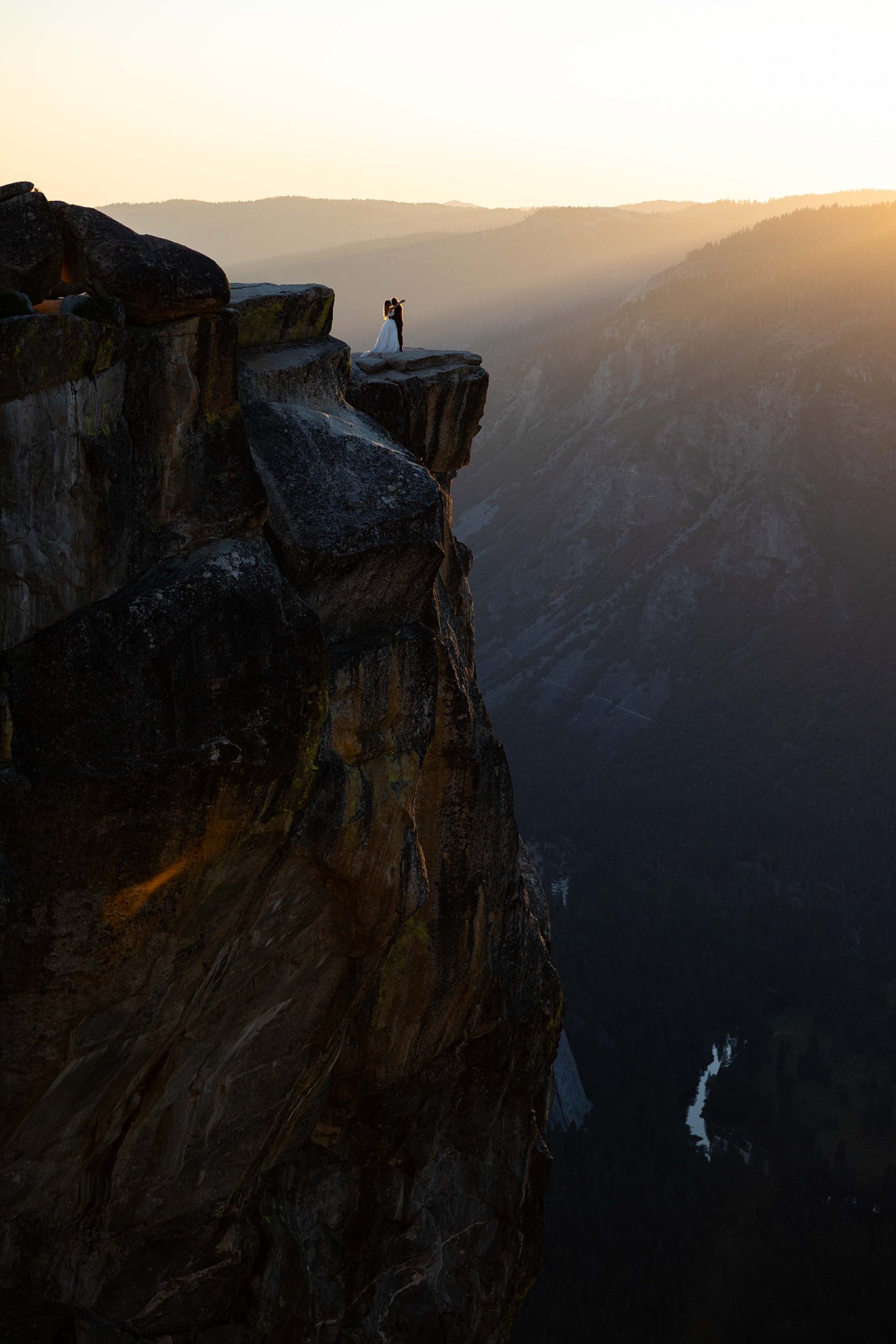 A Bride and Groom stands on the edge of Taft Point in Yosemite National Park, bathed in golden sunset light above a dramatic canyon.